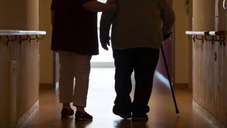 Eine Pflegerin begleitet eine ältere Frau über den Flur eines Pflegeheims.