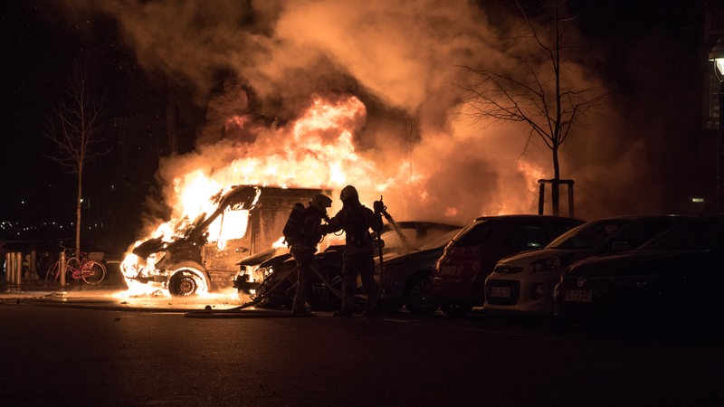 Ein Lieferwagen brennt in einer Straße. Die Feuerwehr löscht ihn.