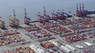 Eine  Luftaufnahme zeigt Containerschiffe die hinter gestapelten Containern am Containerterminal liegen.