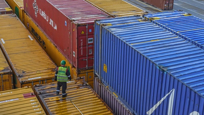 Container in einem Hafen, ein Mann in gelber Wester und mit Helm steht auf einem Container.