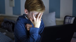 Ein Junge sitzt mit einem Laptop auf seinem Bett, hat die Hand vor das Gesicht geschlagen und schaut sich mit aufgerissenen Augen einen Horrorfilm an.