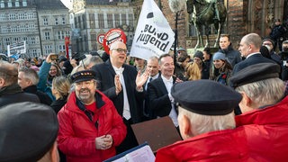 Bremens Bürgermeister und der Bundesverkehrsminister lauschen einem Shantychor. Hinter ihnen stehen Demonstranten.