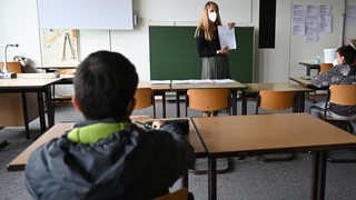Eine Lehrerin steht vor einer Klasse und hält ein Blatt Papier hoch.
