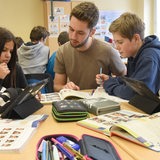 Ein junger Lehrer sitzt mit vier Schülern an einem Tisch und schaut in ein Buch. Zwei Schülerinnen haben Tablets vor sich.