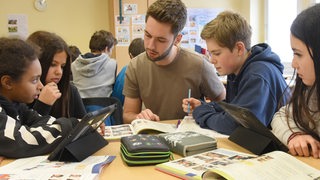 Ein junger Lehrer sitzt mit vier Schülern an einem Tisch und schaut in ein Buch. Zwei Schülerinnen haben Tablets vor sich.
