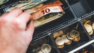 Eine Hand nimmt Geldscheine aus einer Kasse in einem Supermarkt.