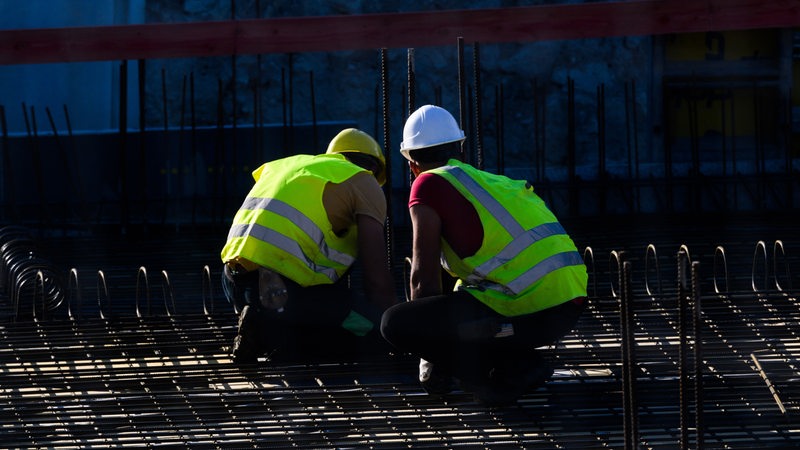 Zwei Männer mit Bauhelmen und Warnwesten hocken auf einer Stahlkonstruktion.