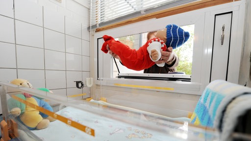 Eine Babypuppe wird durch ein Fenster in ein Bettchen gelegt.