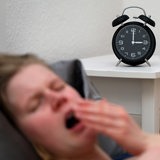 Eine Frau liegt im Bett und gähnt. Die Uhr wird nachts um 2 Uhr auf 3 Uhr vorgestellt.