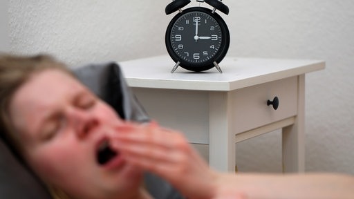 Eine Frau liegt im Bett und gähnt. Die Uhr wird nachts um 2 Uhr auf 3 Uhr vorgestellt.