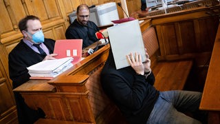 Ein Angeklagter sitzt mit seinen Anwälten im Gerichtssaal und verdeckt sein Gesicht.