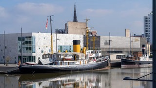 Ein Dampfschiff liegt in einem Hafenbecken vor Gebäuden.