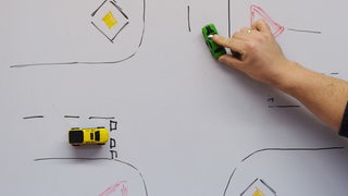Ein Fahrschullehrer erklärt mit Spielzeugautos auf einer Tafel Verkehrsregeln