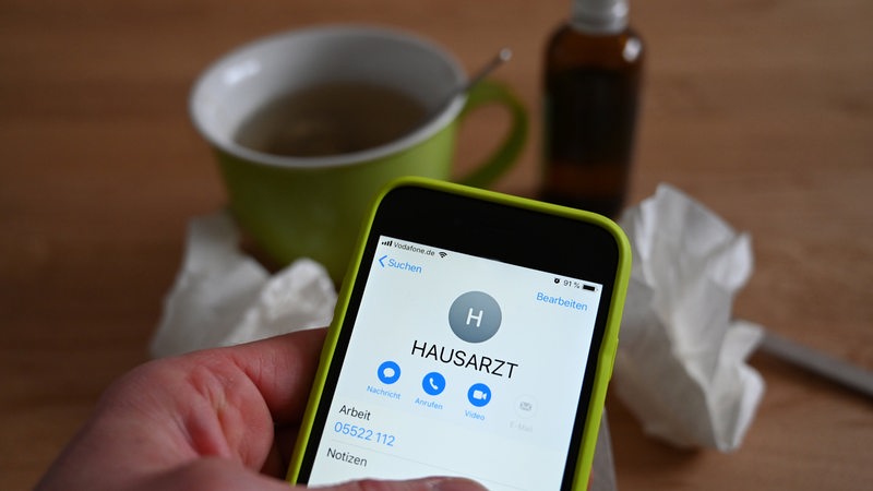Finger wählt Hausarzt-Nummer auf Smartphone, Teetasse und Taschentuch im Hintergrund