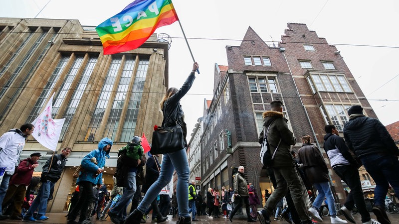 Eine Teilnehmerin des Ostermarsches 2018 geht mit anderen Demonstranten durch die Innenstadt und hält eine Flagge mit der Aufschrift "PACE" nach oben