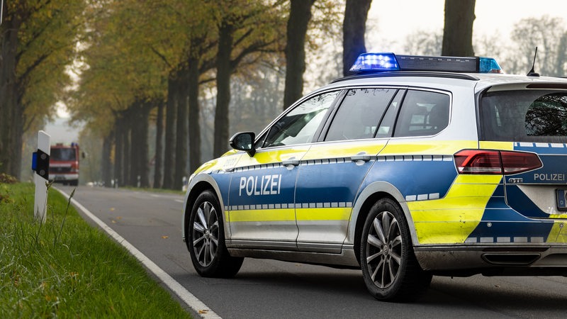 Ein Polizeiauto steht auf einer Landstraße in Niedersachsen. Die Straße ist von Bäumen gesäumt und im Hintergrund ist ein Feuerwehrauto zu sehen.