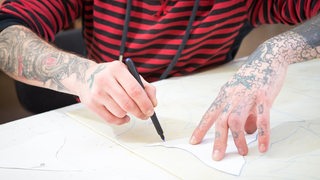 Zwei tätowierte Arme zeichnen etwas auf ein weißes Blatt.