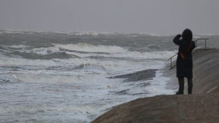 Eine Spaziergängerin macht auf der Insel Norderney bei stürmischem Wetter von der Wasserkante aus Fotos mit einem Handy. 