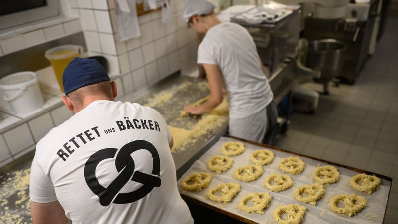 Ein Bäcker und eine Auszubildende stehen am frühen Morgen mit einem T-Shirt „Rettet uns Bäcker“ in der Backstube