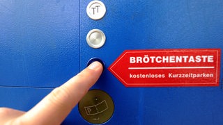 Ein Finger drückt auf einem Parkscheinautomat eine Taste, die mit "Brötchentaste" beschriftet ist.