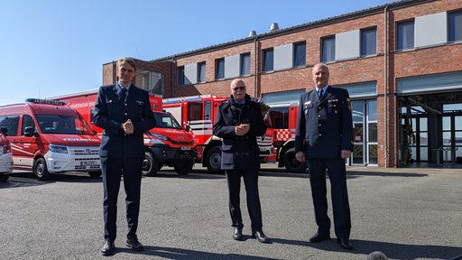 Der Chef der Bremer Feuerwehr, der Bremer Innensenator und ein weiterer Mann stehen vor Feuerwehrautos.