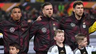 Die Nationalspieler Thilo Kehrer, Niclas Füllkrug und Leon Goretzka singen. Vor ihnen stehen Kinder in DFB-Trikots.
