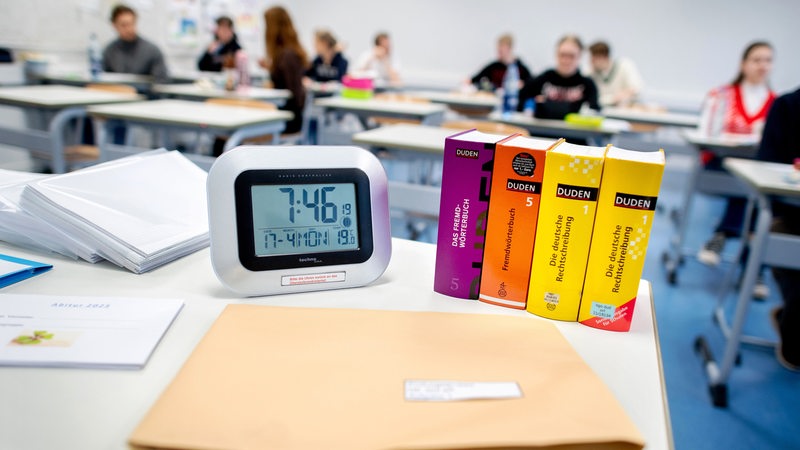 Eine digitale Uhr und mehrere Wörterbücher stehen vor Beginn einer schriftlichen Abiturprüfungen auf einem Tisch.
