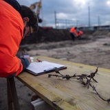 Ein Mann auf einer Baustelle schreibt etwas. Auf dem Tisch neben ihm liegt alter Stacheldraht.