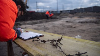 Ein Mann auf einer Baustelle schreibt etwas. Auf dem Tisch neben ihm liegt alter Stacheldraht.