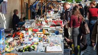 Besucher eines Flohmarkts stehen an einem Verkaufstisch. 