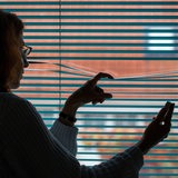 Eine Frau beobachtet durch eine geschlossene Jalousie nach einem Stalking-Anruf die Straße vor ihrem Haus