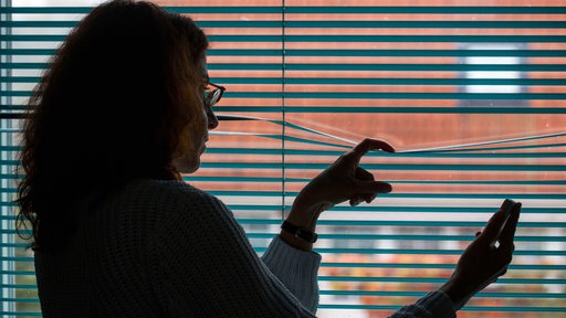 Eine Frau beobachtet durch eine geschlossene Jalousie nach einem Stalking-Anruf die Straße vor ihrem Haus