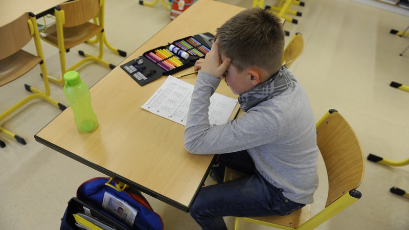 Ein Schüler sitzt bei einer Klassenarbeit am Tisch und stützt mit der Hand nachdenkend den Kopf.