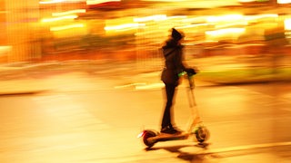 Eine junge Frau fährt nachts auf einem E-Tretroller auf einer Straße.