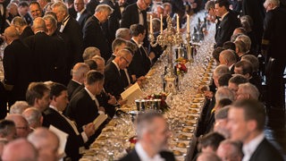 Männer sitzen an einem langen festlich gedeckten Tisch mit Gläsern und Kerzenständern. Die Gäste tragen dunkle Anzüge. 