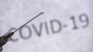 Eine Spritze, an der ein Tropfen hängt, im Hintergrund ein Schriftzug "Covid-19"