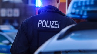 Ein Polizist steht in der Dämmerung neben einem Polizeiauto. Im Hintergrund blinkt Blaulicht.