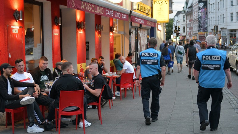 Zwei Polizisten mit blauen Westen mit der Aufschrift "Kommunikationsteam" laufen im Bremer Viertel eine Straße entlang. Leute sitzen auf Stühlen vor einem Lokal.
