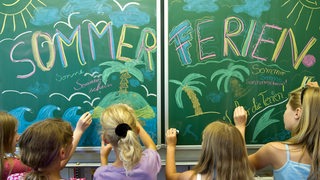 Schülerinnen einer 5. Klasse gestalten vor der Zeugnisausgabe und dem Beginn der Sommerferien eine Schultafel in ihrem Klassenraum mit dem Wort "Sommerferien".