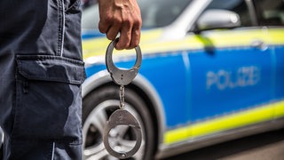 Ein Polizist mit Handschellen in der Hand steht vor einem Polizeiauto.