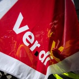 Eine Person in Warnweste ist durch eine rote Fahne mit der Aufschrift ver.di zu sehen.