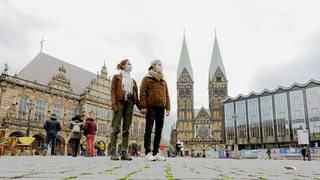 Ein Mann und eine Frau stehen vor dem Alten Rathaus, dem Bremer St.-Petri-Dom und der Bremischen Bürgerschaft und tragen Mund-Nasen-Schutz.