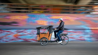 Ein Mann mit Helm fährt mit einem Lastenrad an einem Graffiti vorbei.