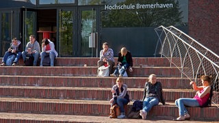 Auf einer Treppe zu einem Gebäude der Hochschule Bremerhaven sitzen Menschen in der Sonne.