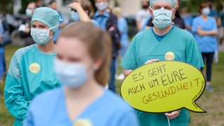 Eine Pflegefachkraft mit Maske, im Hintergrund ein Schild mit der Aufschrift "Es geht auch um Eure Gesundheit"
