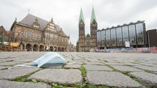 Ein Mund-Nasen-Schutz liegt auf den Pflastersteinen vor dem Alten Rathaus, dem Bremer St.-Petri-Dom und der Bremischen Bürgerschaft. 