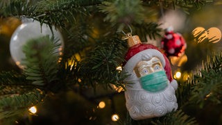 An einem Weihnachtsbaum hängt Weihnachtsschmuck in Form eines Weihnachtsmannes, der eine Maske trägt (Symbolbild)