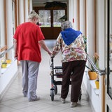 Eine Pflegerin geht mit einer Heimbewohnerin am Rollator durch den Flur eines Pflegeheims.