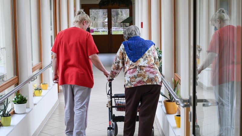 Eine Pflegerin geht mit einer Heimbewohnerin am Rollator durch den Flur eines Pflegeheims.