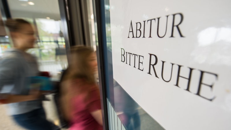 Auf einem Schild steht "Abitur, bitte Ruhe!" Dahinter gehen Schüler durch eine Tür.